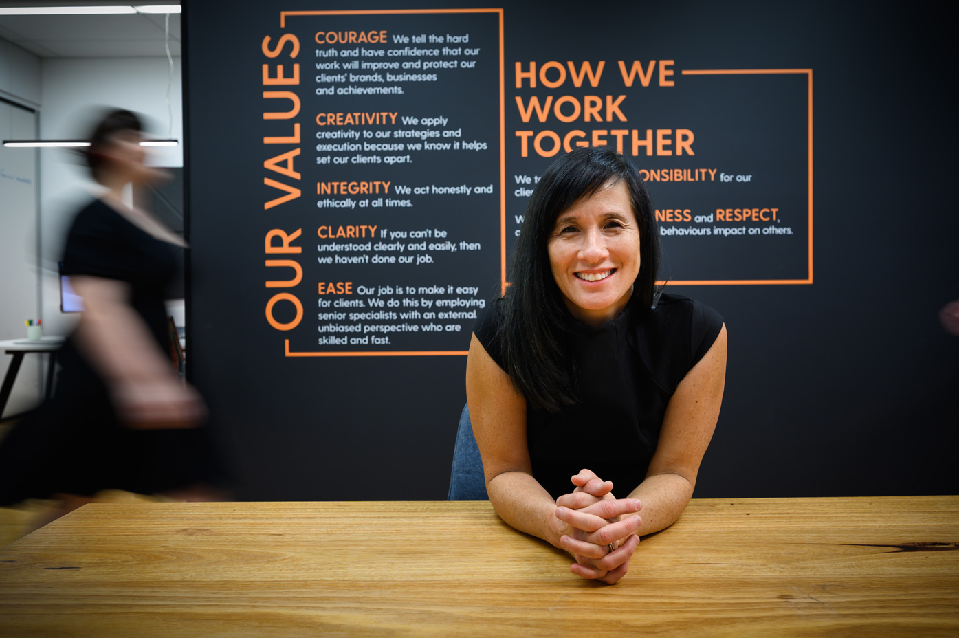 Amanda Newbery sitting at a desk smiling with her hands folded. Behind her is a dark wall with a decal displaying 'our values' and 'how we work together'