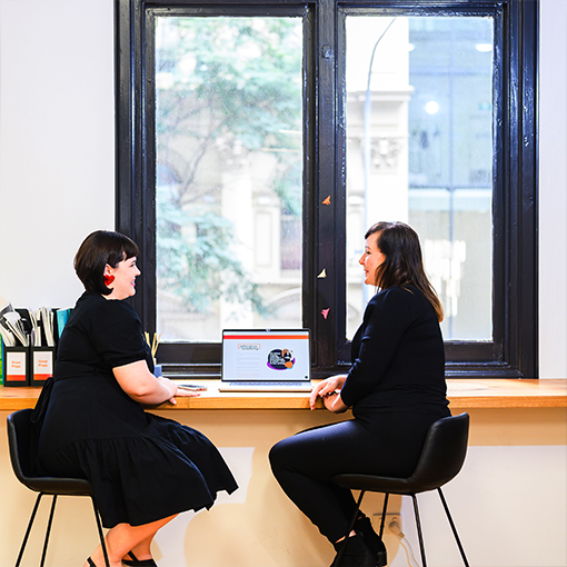 Two ladies sitting on stools at a work bench with a window, in front of a laptop, talking to each other