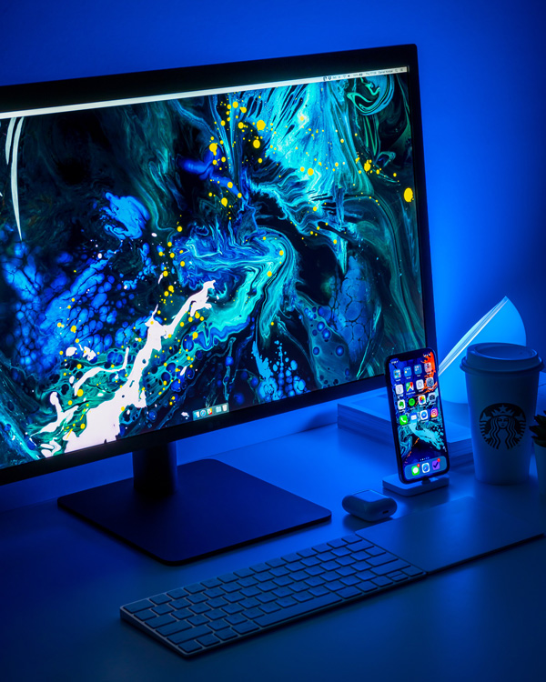 An image of a workstation with a desktop computer with a blue paint texture wallpaper and an iPhone in a blue neon lighting background