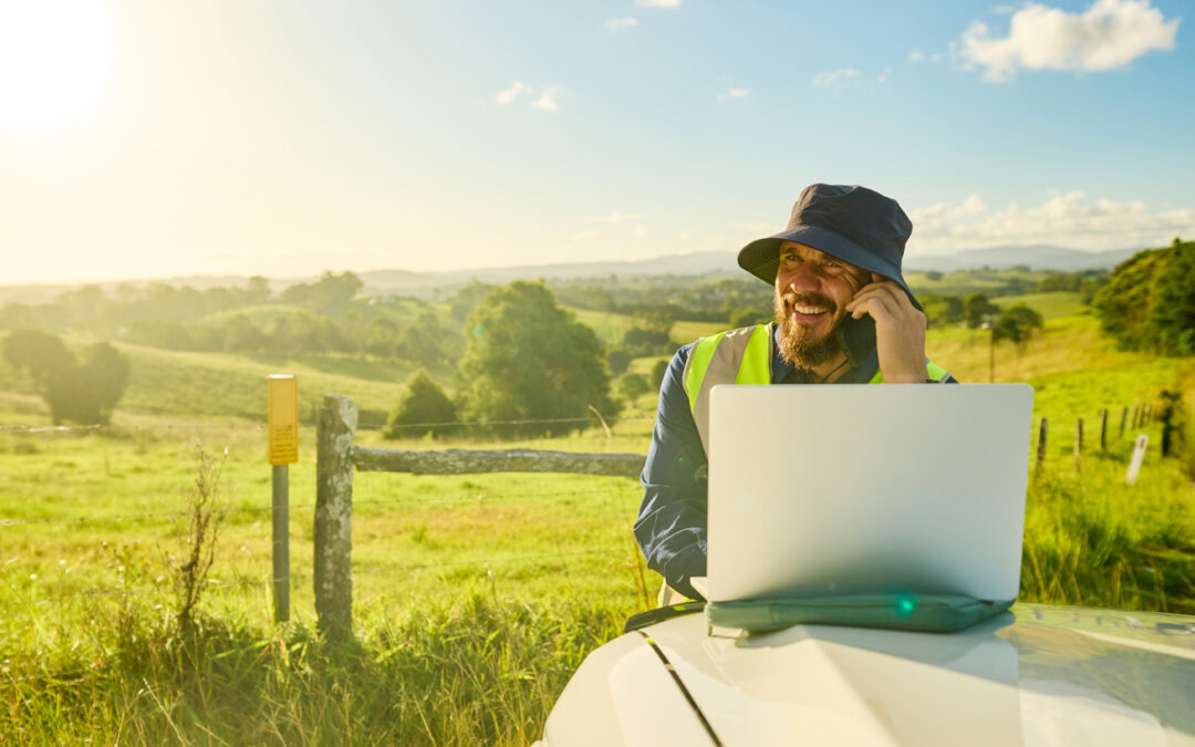 Smiling engineer wearing high-visibility shirt and hat using laptop and phone in a rural landscape, planning a construction project in australia