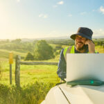 Smiling engineer wearing high-visibility shirt and hat using laptop and phone in a rural landscape, planning a construction project in australia
