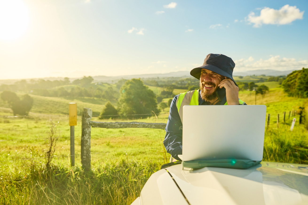 Smiling engineer wearing high-visibility shirt and hat using laptop and phone in a rural landscape, planning a construction project in australia